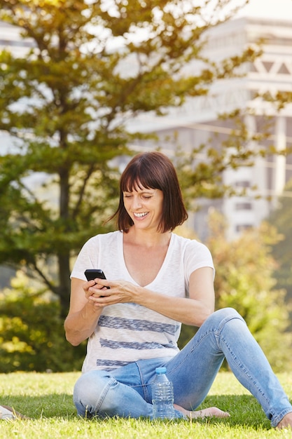 Donna felice che si siede nell&#39;erba con acqua con il cellulare