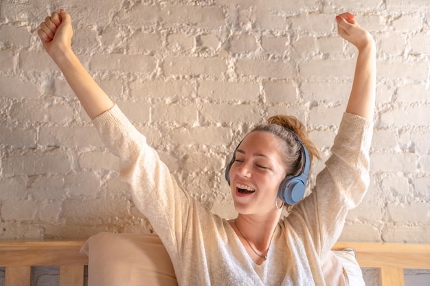 Donna felice che si siede sul letto e che ascolta musica in cuffia divertendosi. Sorridere e ballare.