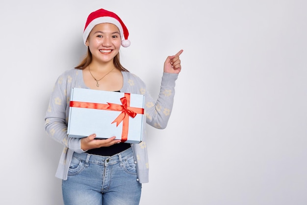 Donna felice che tiene in mano un regalo di Natale Bella donna asiatica in cappello di Babbo Natale che punta il dito contro lo spazio della copia isolato su sfondo bianco per studio Merry Christmas Concept