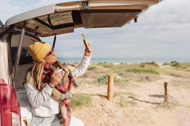 Donna felice che tiene una tazza appoggiata a un furgone vicino alla spiaggia e si fa un selfie