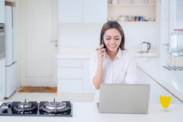 Donna felice che usa il cellulare e il computer portatile in cucina