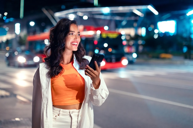 Donna felice che usa lo smartphone guardando da parte e sorridendo camminando sulla strada notturna della città piena di neon