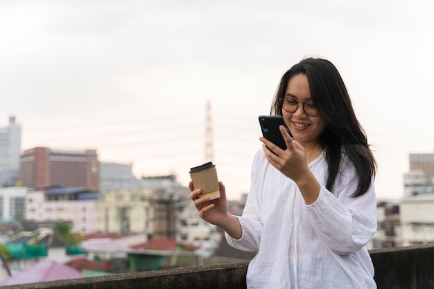 Donna felice che utilizza il telefono cellulare sorridente sul tetto Utilizzo di uno smartphone durante una pausa rilassante con il caffè