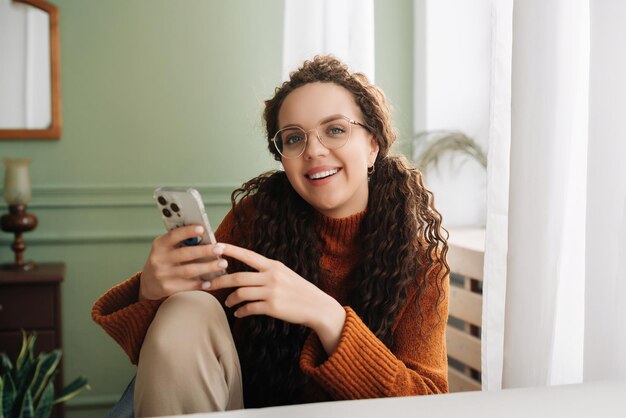 Donna felice che utilizza la tecnologia del telefono cellulare a casa sorridendo mentre controlla le app sul suo smartphone e si rilassa con i dispositivi cellulari in mano