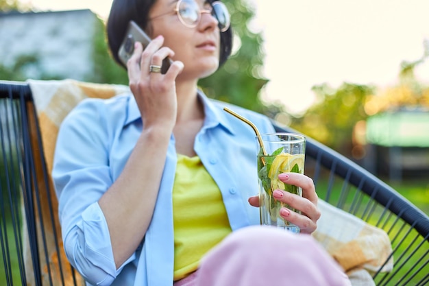 Donna felice con gli occhiali che parla al telefono cellulare seduto sulla sedia al cortile all'aperto