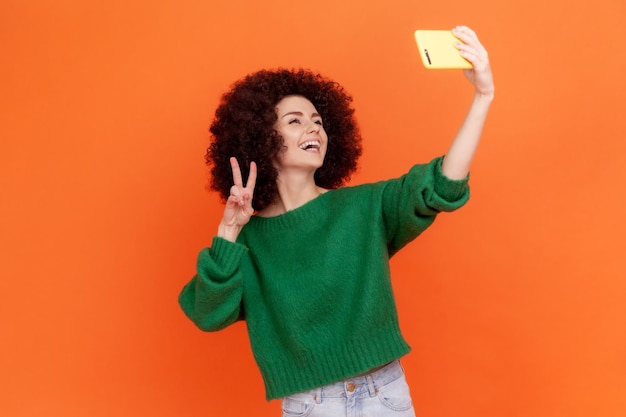 Donna felice con l'acconciatura afro che indossa un maglione verde in stile casual che ha una conversazione online con i follower nei social network che mostrano il segno v Indoor girato in studio isolato su sfondo arancione