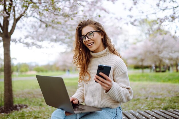 Donna felice con laptop e telefono nel parco in fiore di primavera mentre lavora online concetto di freelance