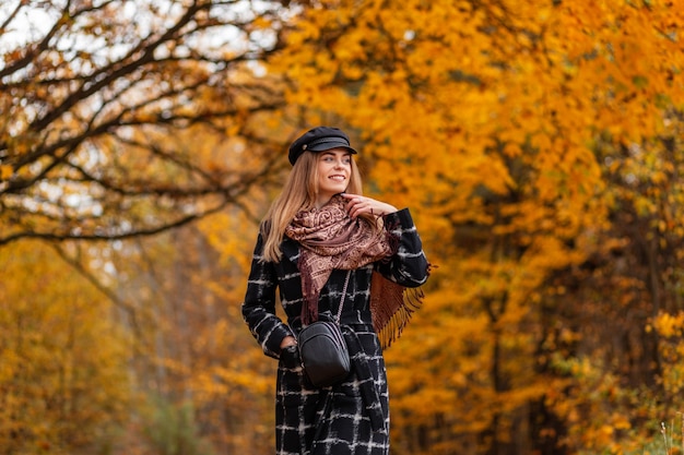 Donna felice con un sorriso carino in cappotto alla moda, sciarpa e cappello cammina nel parco autunnale con fogliame autunnale dorato