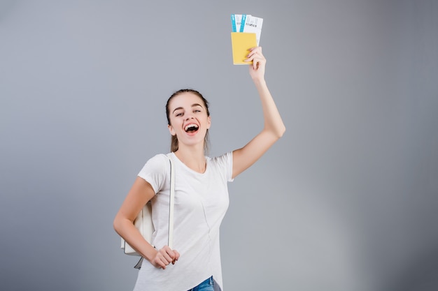 Donna felice del brunette con i biglietti di aeroplano in passaporto e zaino isolati sopra grey