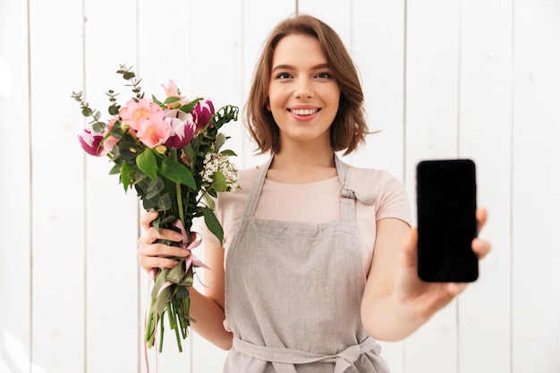 Donna felice del fiorista che sta con i fiori che mostrano esposizione del telefono cellulare.