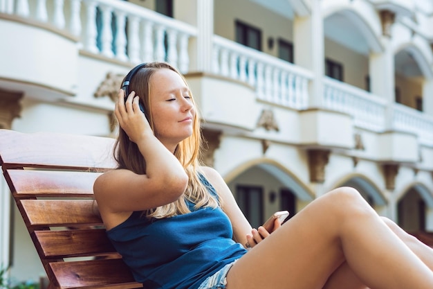 Donna felice dello smartphone che si rilassa vicino alla piscina ascoltando con gli auricolari la musica in streaming. Bella ragazza che utilizza i dati 4g dell'app del suo telefono cellulare per riprodurre brani mentre si rilassa durante le vacanze estive di lusso