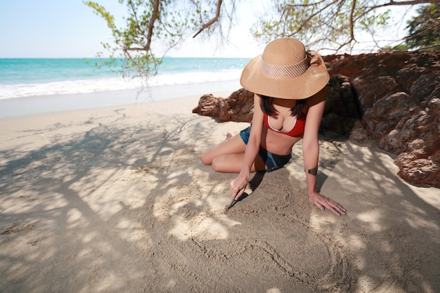 Donna felice e sexy con il simbolo del cuore del disegno del cappello sulla spiaggia sabbiosa