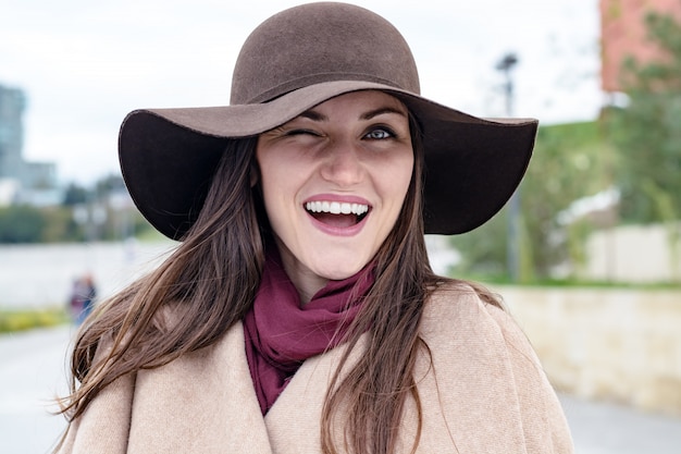 Donna felice in cappello marrone e cappotto beige, facendo l'occhiolino di un occhio alla macchina fotografica e sorride ampiamente con denti bianchi.e.