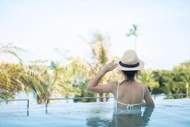 Donna felice in costume da bagno bianco che nuota in un hotel di lusso con piscina giovane femmina con cappello godere in resort tropicale Rilassante vacanza estiva e concetto di fine settimana