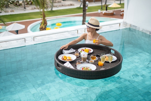 Donna felice in costume da bagno bianco con vassoio colazione galleggiante in hotel di lusso con piscina