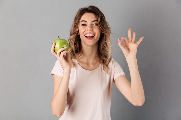 Donna felice in maglietta che fora mela fresca e che mostra segno giusto mentre sopra gray