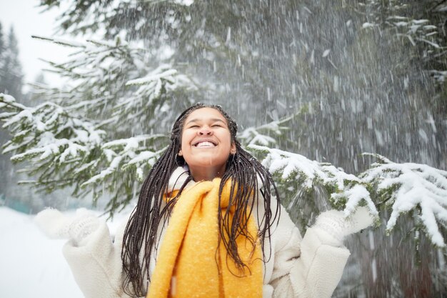 Donna felice in sciarpa calda in piedi sotto la neve che cade dagli alberi nella foresta