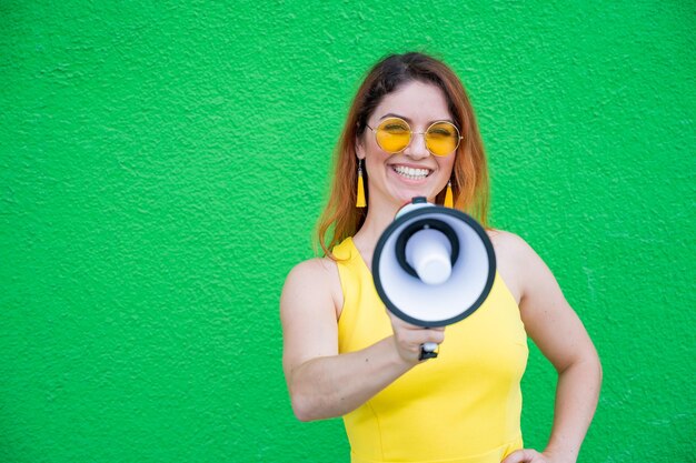 Donna felice in un abito giallo occhiali e orecchini su sfondo verde che grida in un megafono Ritratto di una ragazza con un altoparlante Signora con un perfetto sorriso bianco come la neve