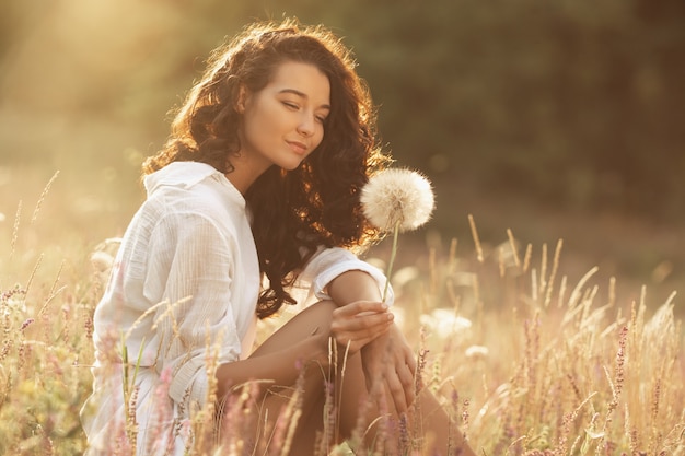 Donna felice libera che gode della natura. Ragazza di bellezza all'aperto. Concetto di libertà. Ragazza di bellezza con il dente di leone, giacimento di fiori soleggiato. Raggi di sole. Piacere e tenerezza. Foto ritratto