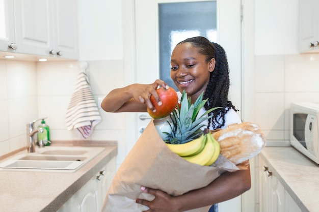 Donna felice positiva sana che tiene un sacchetto della spesa di carta pieno di frutta e verdura. Giovane donna che tiene la borsa della spesa con verdure in piedi in cucina