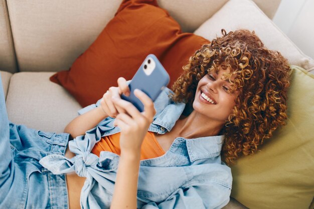 Donna felice seduta sul comodo divano con uno smartphone in mano e sorridente mentre si diverte a giocare online