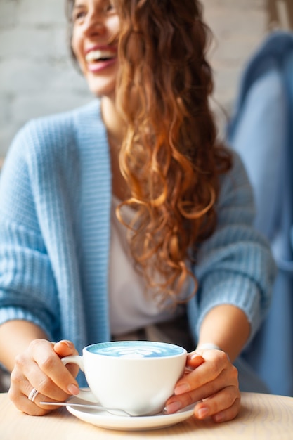 Donna felice sfocata con capelli ondulati lunghi in maglione caldo che tiene tazza di latte blu caldo. Bevanda e colore alla moda. Il latte del tè blu è fatto con fiori di pisello farfalla. Bevanda salutare a base di erbe