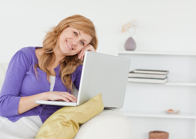 Donna felice sorridente che si siede sul sofà e che per mezzo di un computer portatile