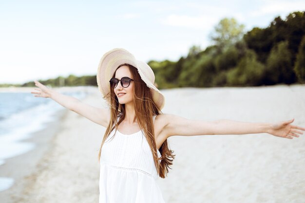 Donna felice sorridente in felicità libera sulla spiaggia dell'oceano in piedi con un cappello, occhiali da sole e mani aperte. Ritratto di una modella femminile multiculturale in abito bianco estivo che si gode la natura durante il viaggio