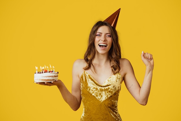 Donna felicissima in abito e cappello di compleanno che tiene la torta posa isolata su sfondo giallo studio