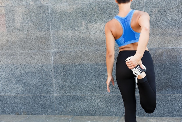 Donna fitness che allunga le gambe prima di correre all'aperto, vista posteriore. La ragazza magra irriconoscibile fa esercizi di aerobica, sfondo grigio della parete, spazio per la copia