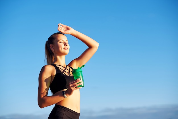 Donna fitness con bottiglia d'acqua dopo aver eseguito l'allenamento sullo sfondo del cielo blu