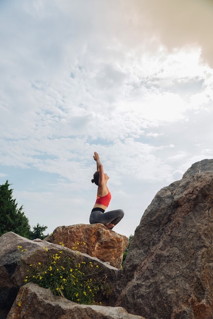 Donna flessibile e sottile, alzando le mani mentre si fa yoga seduto sulla roccia intorno alla natura