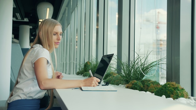 Donna formale con laptop e documenti. Vista laterale di scrittura della donna di affari sulla carta mentre sedendosi con il computer portatile alla tavola vicino alla finestra in ufficio moderno.