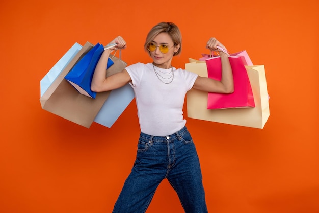 Donna forte e divertente in maglietta bianca e jeans con le braccia alzate con le borse della spesa isolate sullo sfondo arancione