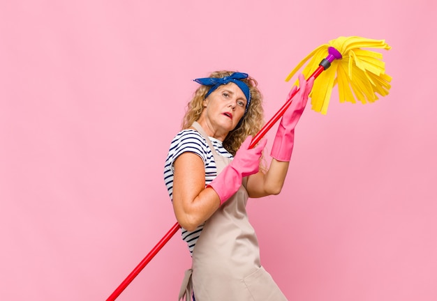 Donna fredda di mezza età con una scopa contro il muro rosa