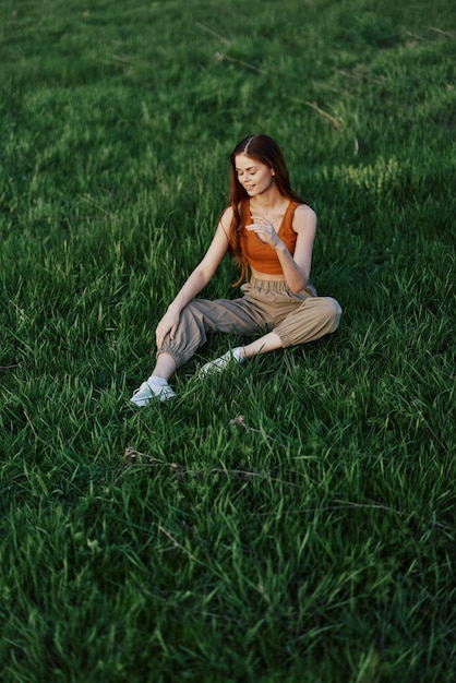 Donna freelance che si gode la vita all'aria aperta seduta nel parco sull'erba verde in abbigliamento casual con lunghi capelli rossi illuminati dal luminoso sole estivo senza zanzare