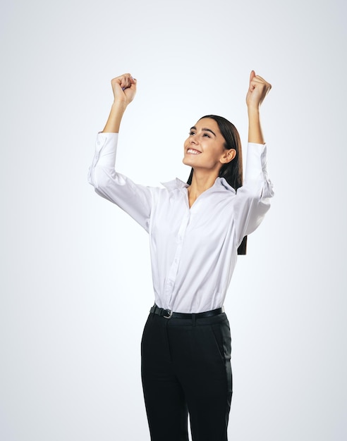 Donna gioiosa e soddisfatta in camicia bianca con le mani alzate che mostrano il suo vincitore del potere e il concetto di successo isolato su sfondo grigio chiaro da vicino