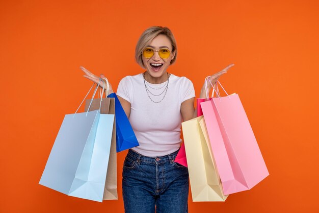 Donna gioiosa in maglietta bianca e jeans in piedi con sacchetti da spesa che si rallegrano isolati sullo sfondo arancione