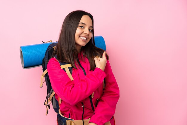 Donna giovane alpinista con un grande zaino sopra la parete rosa isolata che punta indietro