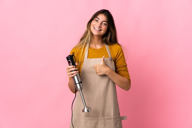 Donna giovane chef utilizzando frullatore a immersione isolato su rosa dando un pollice in alto gesto