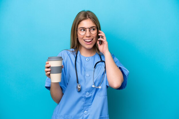 Donna giovane chirurgo medico isolata su sfondo blu che tiene caffè da portare via e un cellulare