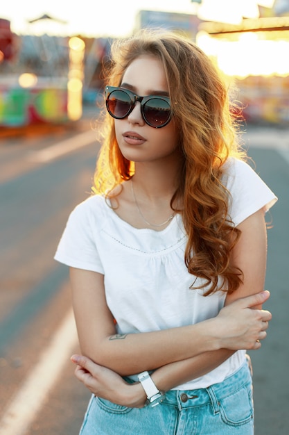 Donna giovane hipster alla moda rossa americana in occhiali da sole alla moda in una t-shirt in jeans vintage in posa