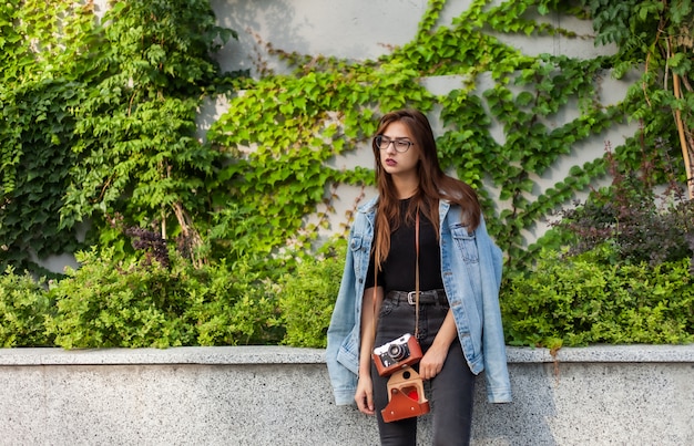 Donna giovane hipster in giacca di jeans, occhiali con pellicola retrò fotocamera all'aperto