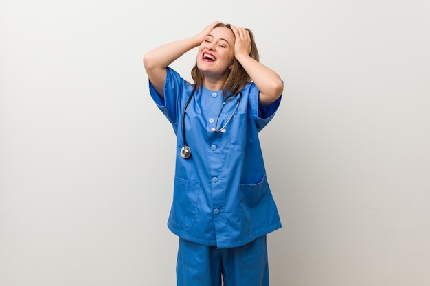 Donna giovane infermiera contro un muro bianco ride con gioia tenendo le mani sulla testa. Concetto di felicità.