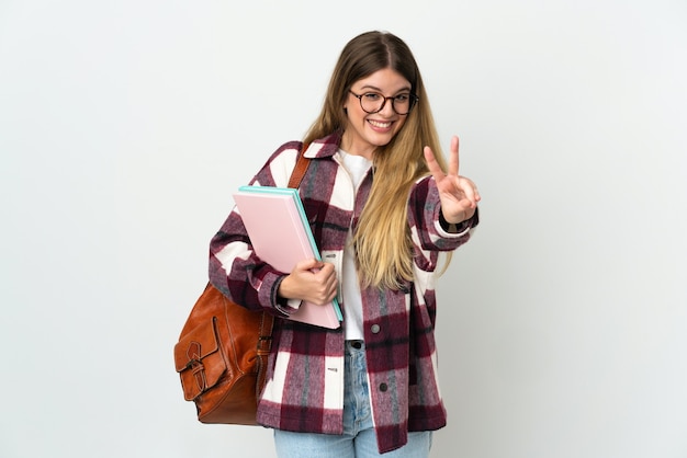 Donna giovane studentessa bionda isolata sul muro bianco sorridendo e mostrando il segno di vittoria