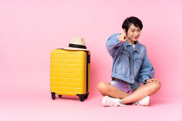 Donna giovane viaggiatore con la valigia che si siede sul pavimento sopra il dito punti rosa con un'espressione sicura