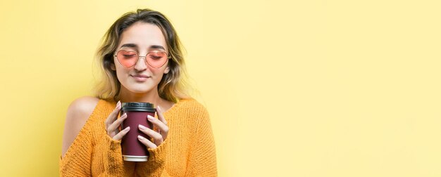 Donna glamour con gli occhiali in un maglione arancione con un drink di caffè su sfondo giallo