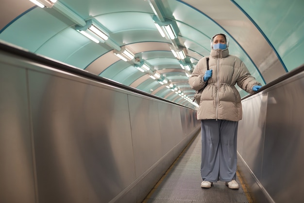 Donna grassoccia in abiti caldi che indossa maschera e guanti a cavallo sulla scala mobile durante la pandemia