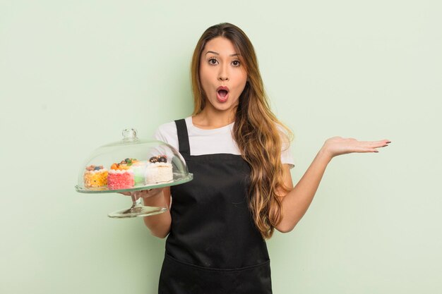 Donna graziosa asiatica che sembra sorpresa e scioccata con la mascella caduta tenendo un concetto di torte fatte in casa di oggetti