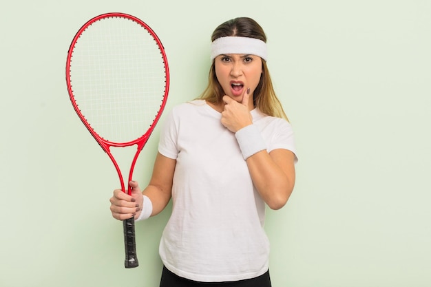 Donna graziosa asiatica con la bocca e gli occhi spalancati e la mano sul concetto di tennis del mento